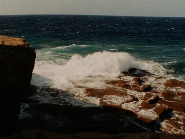Wave crashing on brown rocks, white foam, deep blue ocean
