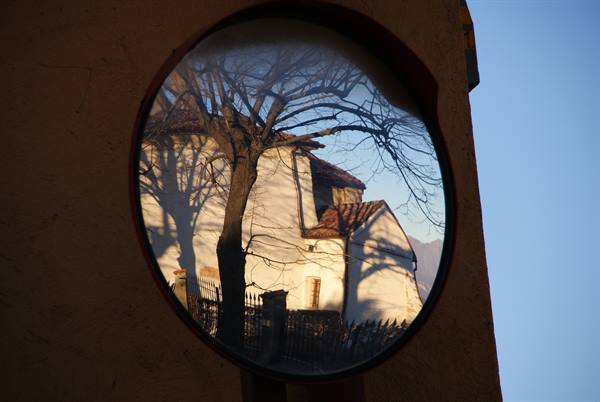 Road mirror reflecting a white house with tiled roof and a bare tree