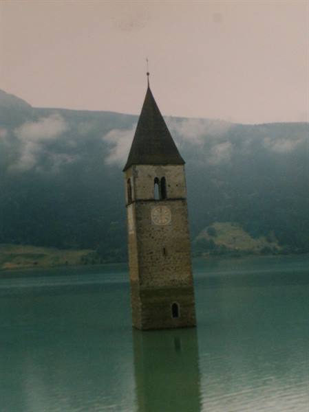 The sunken bell tower of Reschen rises from turquoise water, cloudy mountains behind