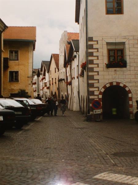 Tourists walk along a cobbled alley past old houses with flower boxes