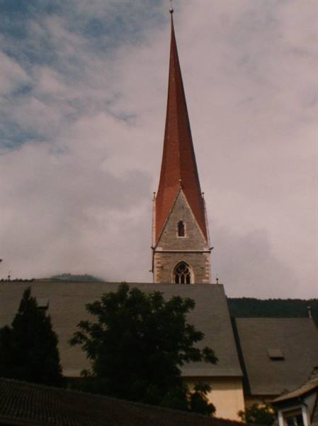 Stone bell tower with red spire above rooftops, trees and clouds