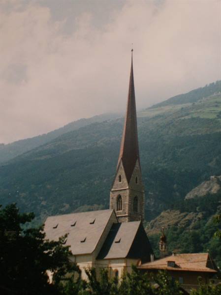 Stone church tower with reddish spire above rooftops, green mountains behind
