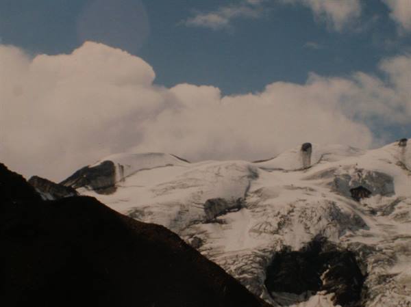 Snowy glacier among dark rocks under white clouds and blue sky