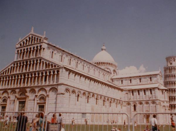 Pisa Cathedral in white marble with tourists behind the fence, leaning tower at right