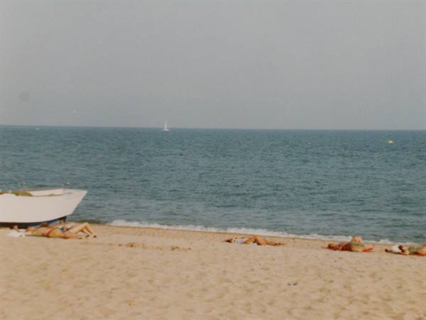 Sunbathers lying on sand, white boat ashore, sailboat in distance