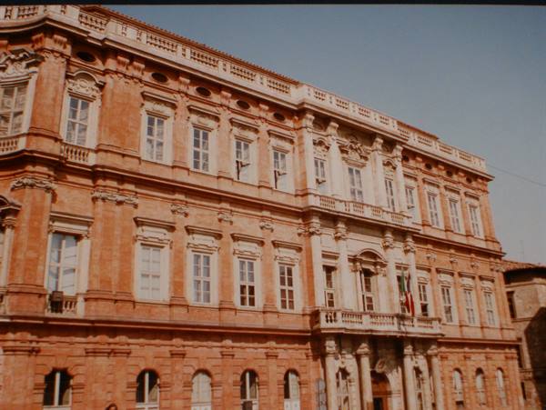 Red-brick baroque palace with tall windows and Italian flag in sunlight