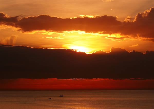 Fiery sun breaking through clouds over the sea, small boats on horizon