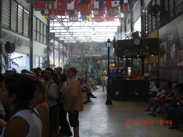Travelers queue under hanging flags inside covered market, Café do Tiém kiosk