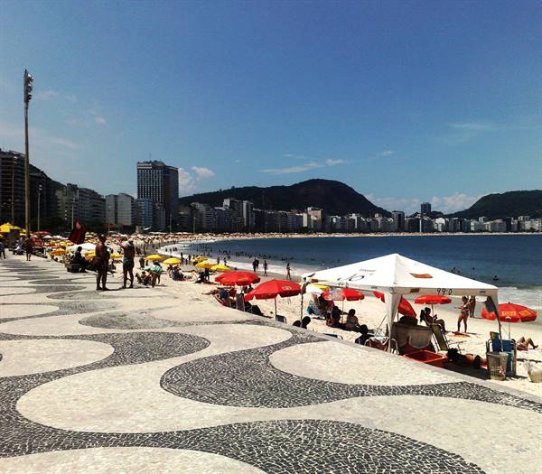 Copacabana's wavy mosaic promenade with red umbrellas and beachgoers under bright sun