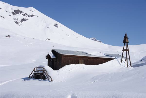 Wooden chapel half-buried in snow among snowy mountains, clear blue sky