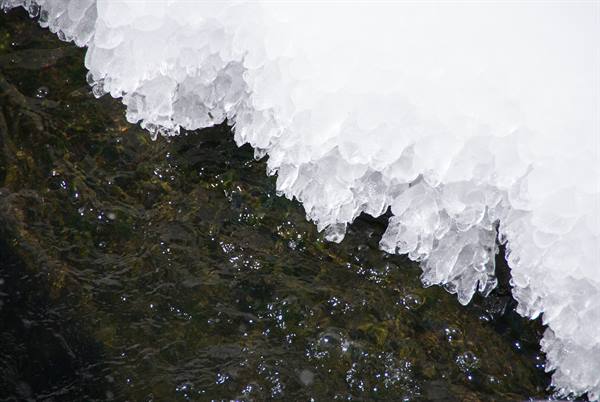 Snowy ice formations hang over dark stream water flowing beneath