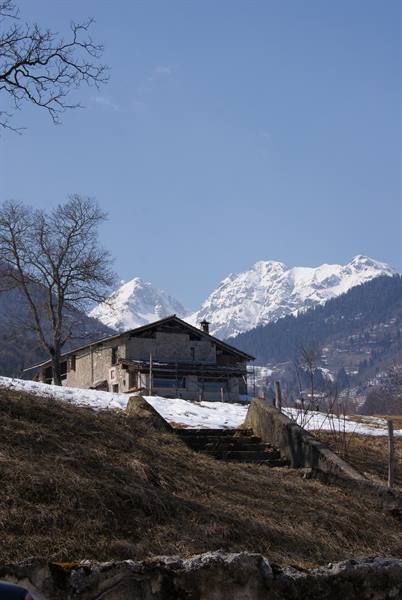 Stone farmhouse with snow, concrete steps, snowy mountains and bare trees