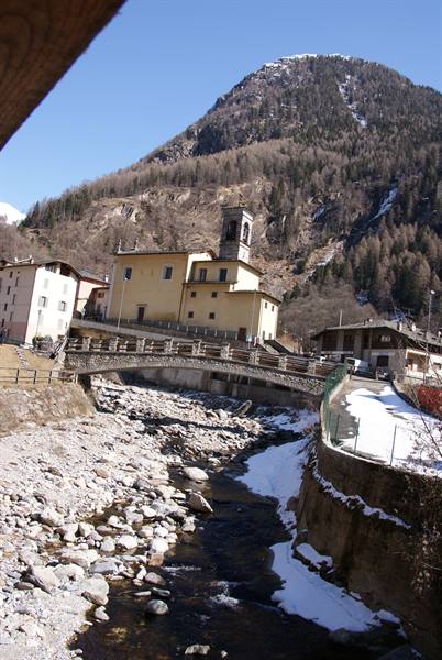 Yellow church with bell tower above stone bridge, rocky stream and snowy mountain