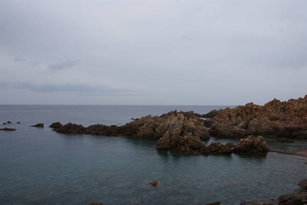 Red granite rocks plunge into a calm sea under a grey sky