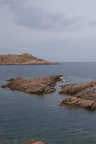 Pink granite rocks in blue water, green rocky hill under cloudy sky