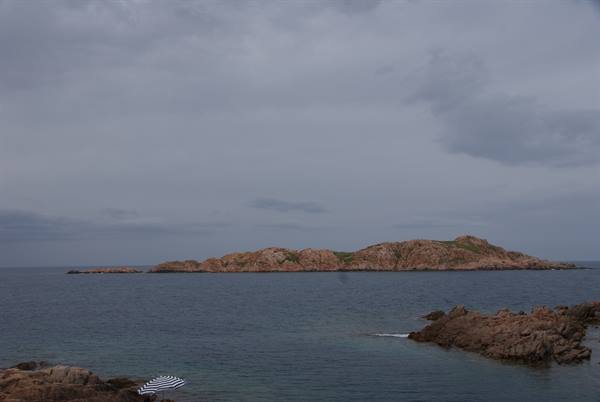 Pink granite islet offshore, striped umbrella on the rocky shore under cloudy sky
