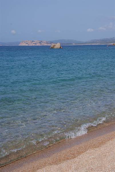 Clear waves on pale sand, rock and reddish coasts in the background