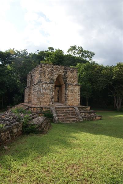 Small Maya stone temple with arched doorway and staircase, surrounded by green trees