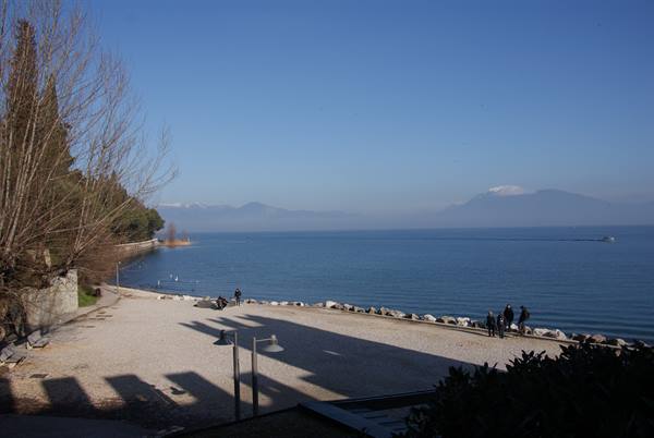 Pebbly lakeside promenade with strolling people and misty mountains, Lake Garda