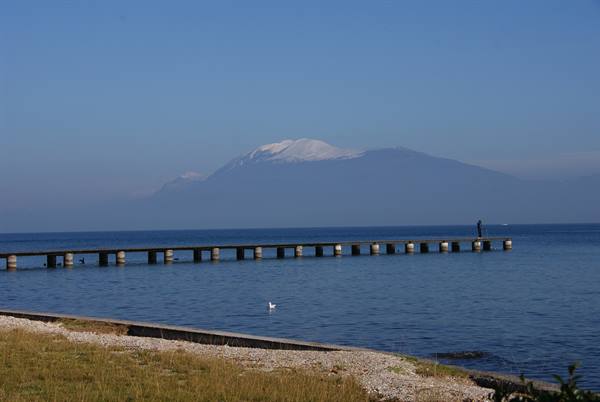 Concrete pier over calm sea, lone figure fishing, snow-capped Mount Athos behind