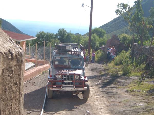 Laden Jeep on dirt road, people nearby, sea and green hills in background