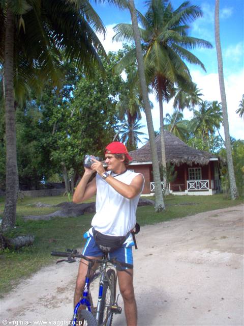 Young man in red cap drinks beside his bicycle among palm trees and thatched bungalow