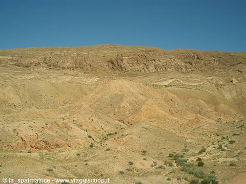 Arid sandy and rocky hills under blue sky, sparse green shrubs