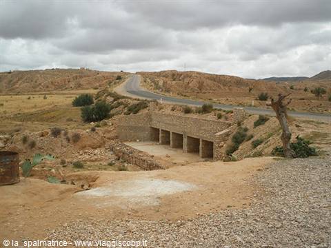 Paved road through arid hills and a stone bridge under gray clouds