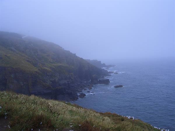 Foggy grassy cliffs above a rocky shoreline, sea breaking on rocks
