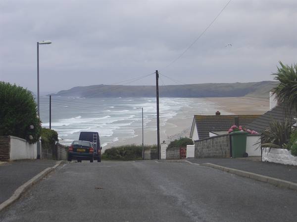 Asphalt road slopes down to the beach, parked car, low houses, grey sky