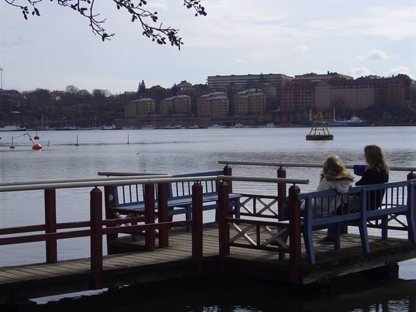 Two women sitting on blue wooden benches on a pier by Stockholm bay