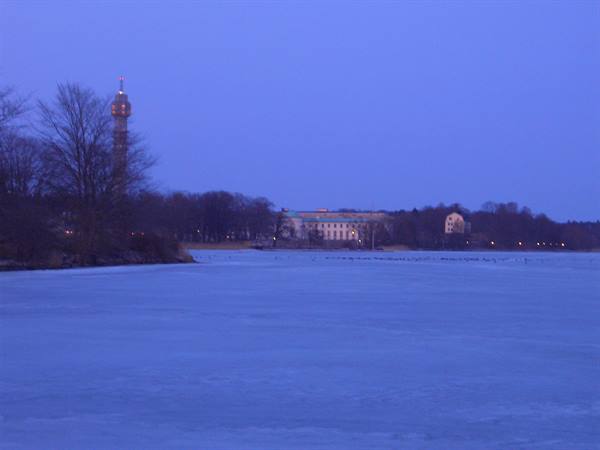 Frozen lake before lit buildings and Kaknästornet tower at dusk