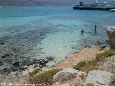 Clear turquoise water, swimmers among rocks, ferry docked in Crete