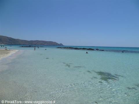 Pink sand beach with turquoise shallow water, low rocks, swimmers, dry hills Elafonissi