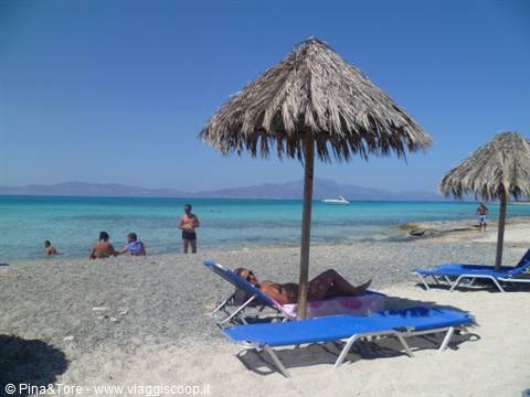 Sandy beach with straw umbrellas and blue sunbeds, swimmers in turquoise water