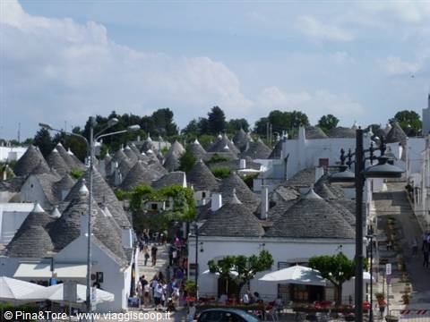 Tourists stroll among Alberobello trulli, conical stone roofs under clouds