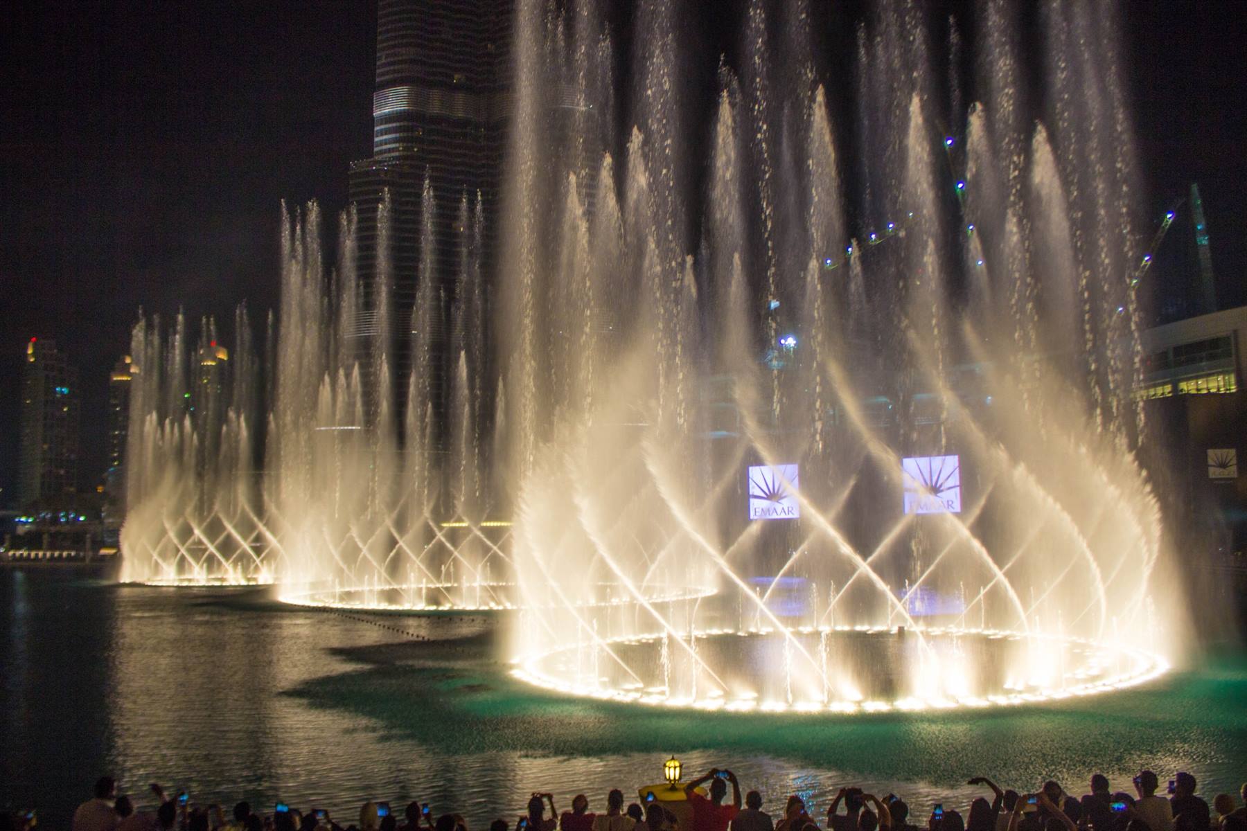 Getti d'acqua illuminati di notte, folla spettatrice lungo la riva del lago