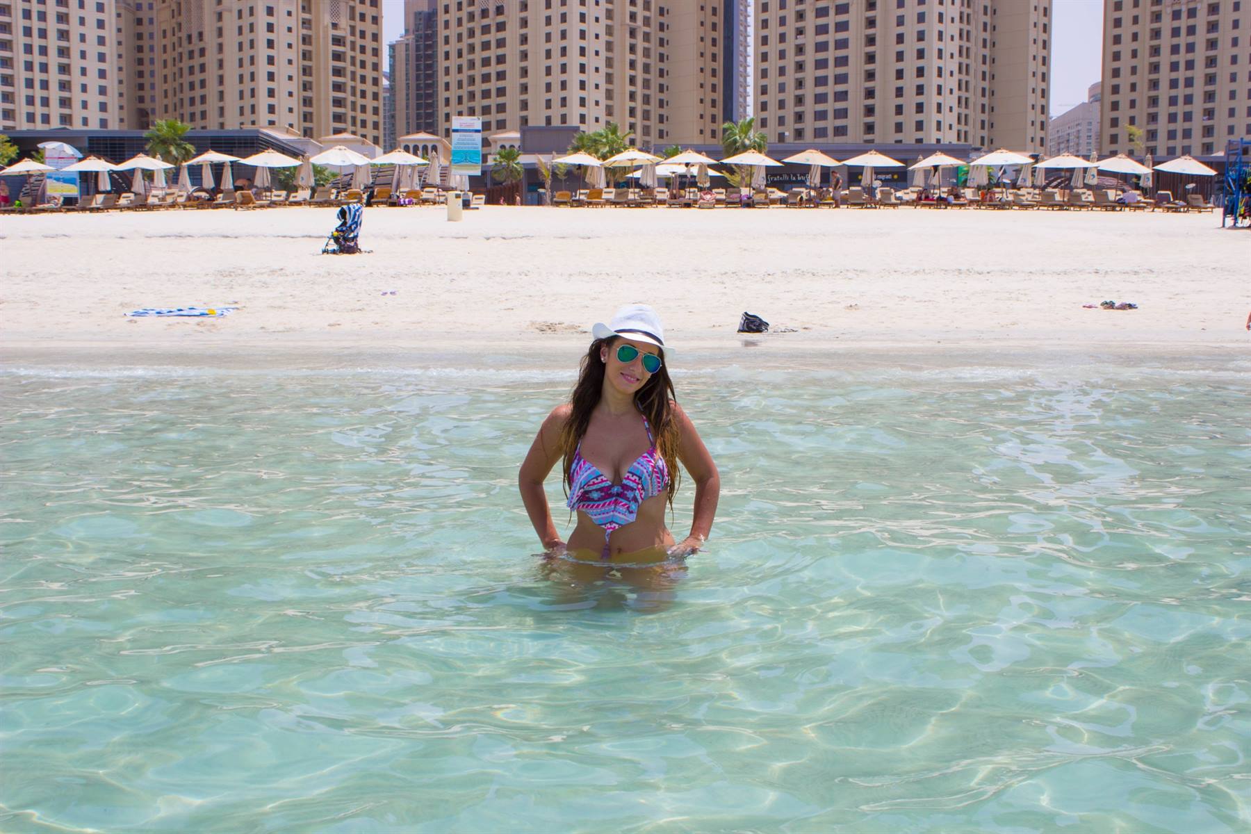 Donna con cappello bianco in mare turchese davanti a spiaggia con ombrelloni e grattacieli