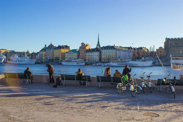 People on benches along Stockholm's waterfront, bicycles and ferries on the bay