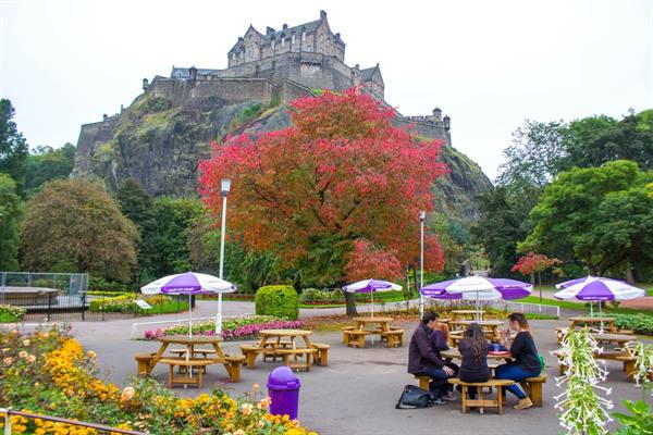 Edinburgh Castle on the rock, wooden picnic tables with umbrellas, tourists sitting, red tree