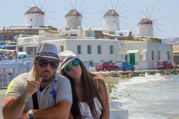 Two tourists in hats in front of Mykonos windmills, white houses and sea