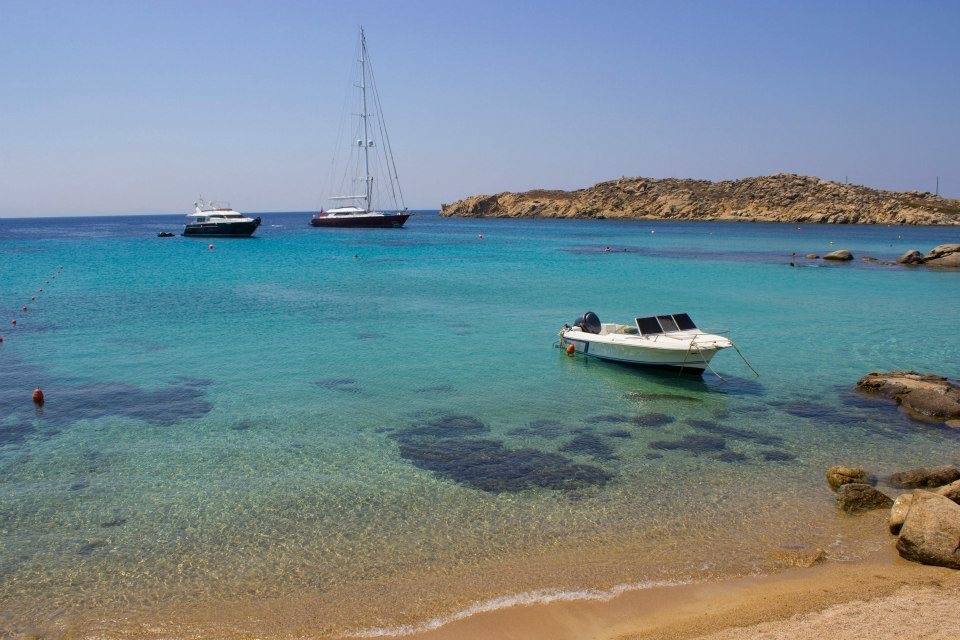 Barche ancorate in baia turchese, scoglio dorato e sabbia chiara sotto cielo azzurro