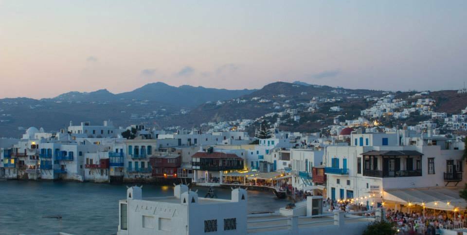 Case bianche sul lungomare di Mykonos, luci accese, montagne al crepuscolo