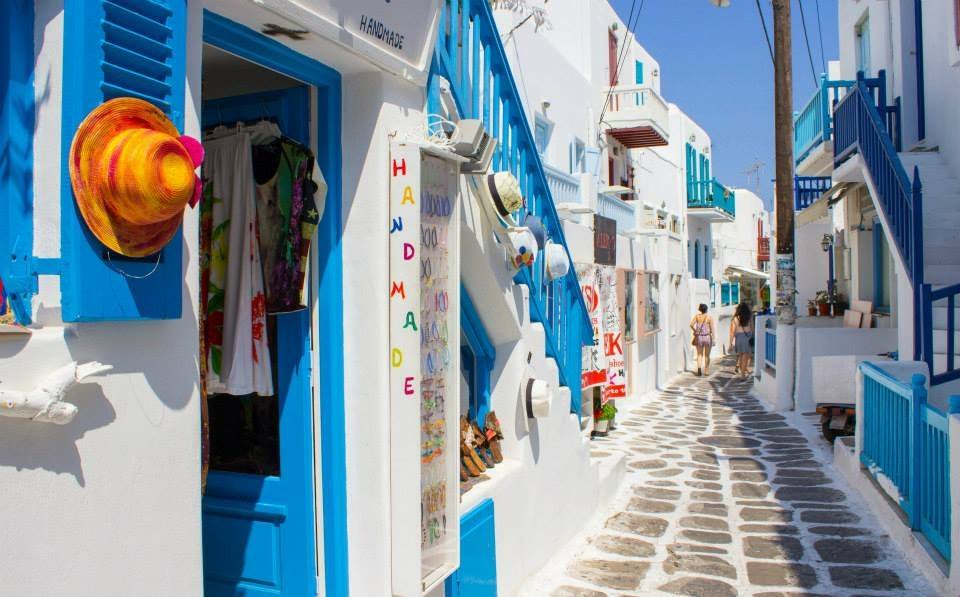 Vicolo di Mykonos con negozi bianchi e porte blu, turisti camminano tra scalinate