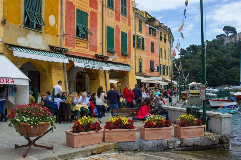 Turisti ai tavolini del bar nel porto di Portofino tra case colorate e fiori
