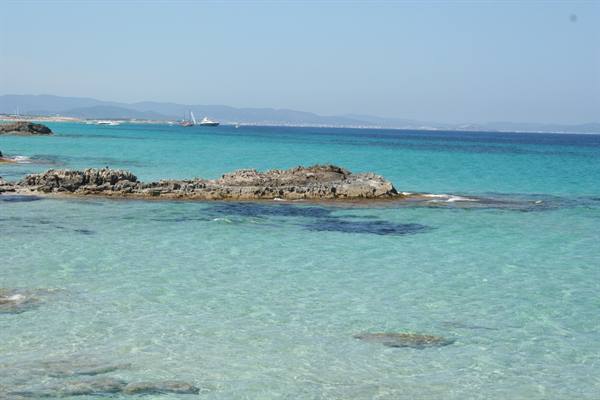 Grey rocks emerging from clear turquoise water, boats and coastline in background