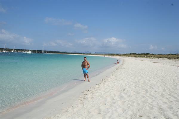 Man in blue swim shorts on white sand, turquoise water with anchored boats