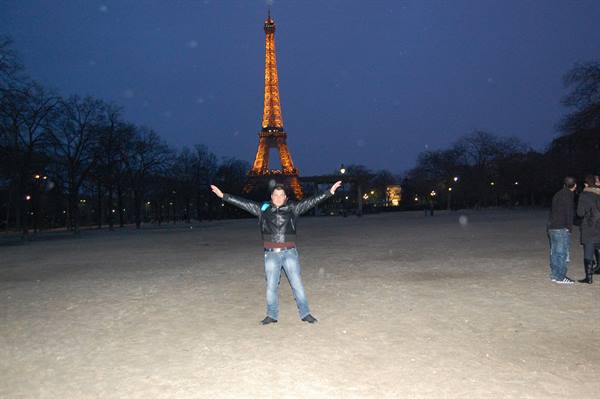 Man in leather jacket arms outstretched in front of the illuminated Eiffel Tower at night