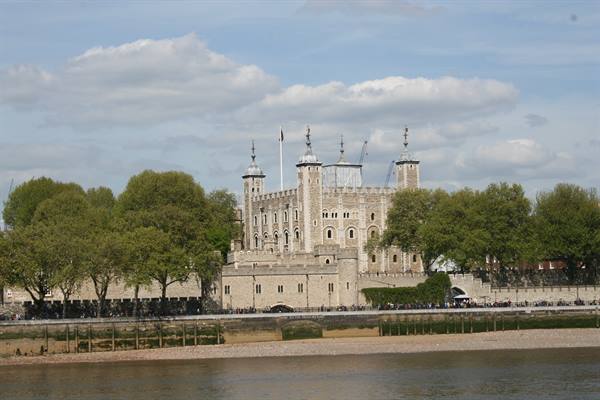 The Tower of London in pale stone among green trees, seen from the Thames under clouds