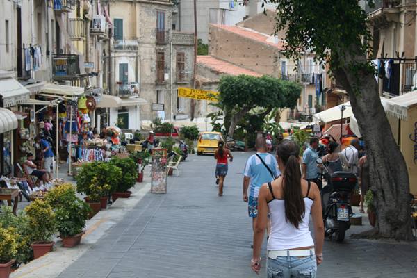 Woman in white top walks down a stone-paved street lined with shops and market stalls